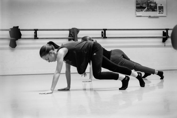 Twee mensen in sportkleding en sokken voeren een vloeroefening uit, geïnspireerd op Hededaagse Dans, in een dansstudio, balancerend op handen en voeten met gebogen knieën. Hun hoofd is gebogen, hun haar is naar achteren gebonden; een barre en jasjes zijn zichtbaar in de zwart-witfoto.