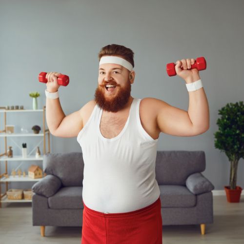 Een lachende man met een baard in een wit tanktopje, rode shorts en een witte hoofdband houdt rode halters vast terwijl hij zich concentreert op het verminderen van visceraal vet tijdens zijn training in de woonkamer. De scène speelt zich af tegen een achtergrond van een grijze bank, decoratieve plank en potplant, wat een vrolijke en casual sfeer uitstraalt.