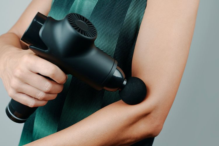 De 10 Beste Massage guns Om Spierpijn Te Verlichten 3 Close-up van een persoon die een zwarte handmassagepistool op zijn bovenarm gebruikt. Het apparaat heeft een ronde, zachte bevestiging die tegen de huid wordt gedrukt. De persoon draagt een mouwloos, groen en zwart gepatroneerd topje. De achtergrond is neutraal grijs.