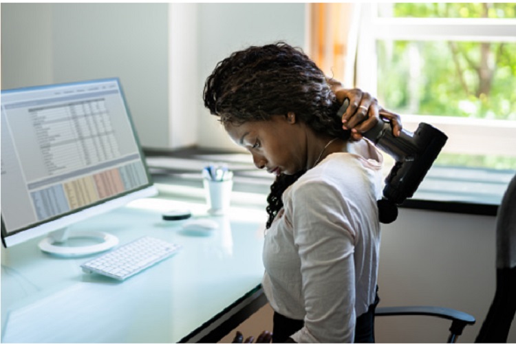 De 10 Beste Massage guns Om Spierpijn Te Verlichten 1 Een vrouw zit aan een bureau en gebruikt een draagbaar massagepistool op haar bovenrug om spierpijn te verlichten. Voor haar toont een computerscherm een spreadsheet. Het bureau bevat ook een toetsenbord, muis en een beker met pennen. Een raam op de achtergrond onthult groen gebladerte buiten.