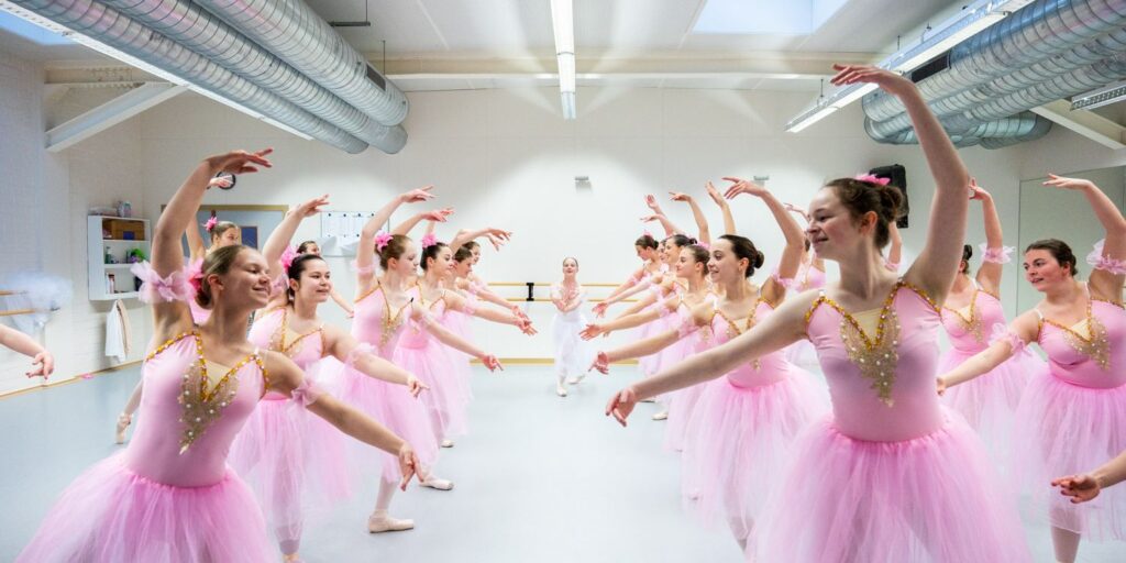 Een groep ballet dansers in roze tutu's en maillots oefent in een felgekleurde dansstudio tijdens een balletl es in Roeselare. Ze staan in twee rijen, met hun armen sierlijk omhoog, glimlachend naar elkaar, terwijl spiegels en balletbarres de muren sieren.