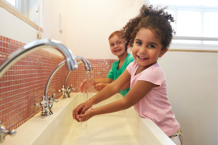 Twee kinderen lachen terwijl ze hun handen wassen bij een wasbak met twee kranen. Het meisje op de voorgrond heeft krullend haar en draagt een roze shirt, terwijl een jongen in een groen shirt naast haar staat. De achtergrond toont een lichtgekleurde muur met een klein betegeld gedeelte in rode en oranje tinten.