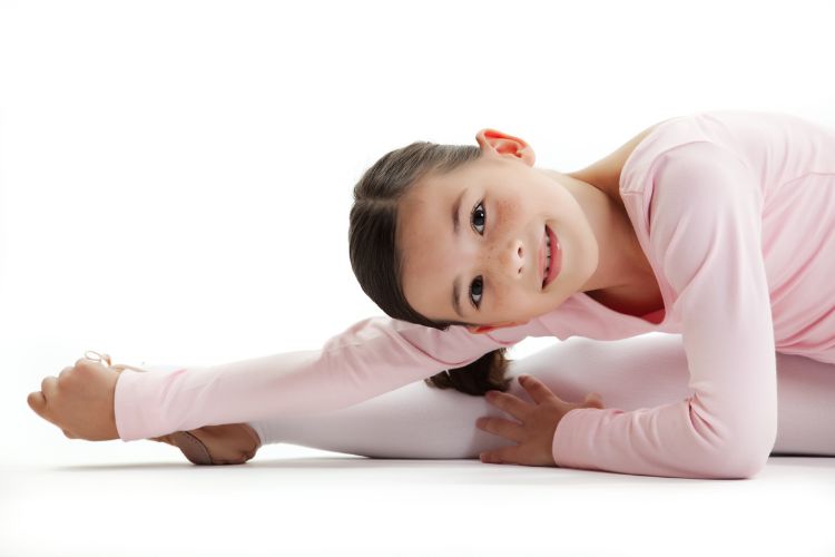 RED-S: Wat het is en hoe je dansers gezond houdt 1 Een jong meisje in een roze balletpakje strekt zich uit op de vloer en lacht naar de camera. Haar rechterbeen is naar voren gestrekt en ze leunt eroverheen, haar voet met beide handen vastpakkend. De achtergrond is helderwit, wat haar sierlijke houding en expressie benadrukt.