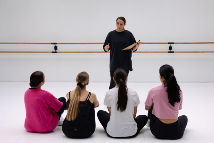 RED-S: Wat het is en hoe je dansers gezond houdt 3 Een dansleraar in een zwarte outfit staat voor vier zittende studenten in een dansstudio met een houten barre aan de muur. De studenten, gekleed in casual sportkleding in roze, zwart en wit, luisteren aandachtig. De kamer heeft een helder, minimalistisch design.