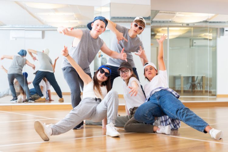 RED-S: Wat het is en hoe je dansers gezond houdt 4 Vijf jongeren gekleed in casual kleding en petten nemen dynamische dansposes aan in een gespiegelde dansstudio. Ze drukken energie en zelfvertrouwen uit, met uitgestrekte armen en glimlachen. De kamer heeft houten vloeren en een muur met grote spiegels die hun bewegingen weerspiegelen.