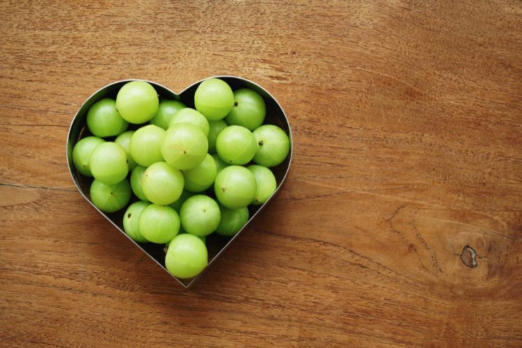 Amla: De Kleine Vrucht met Grote Gezondheidsvoordelen 1 Een hartvormige metalen container gevuld met ronde, groene Indiase kruisbessen wordt op een houten oppervlak geplaatst. Het hout heeft een warme, bruine toon met zichtbare nerven en texturen, wat zorgt voor een natuurlijke en rustieke achtergrond die contrasteert met het levendige groen van de kruisbessen.