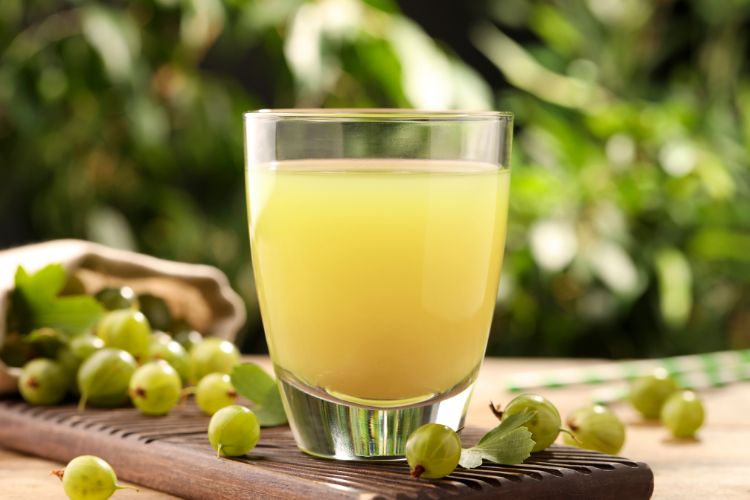 Amla: De Kleine Vrucht met Grote Gezondheidsvoordelen 2 Een glas lichtgroen amla-sap op een houten plank, omringd door verse amla-vruchten met bladeren. De achtergrond is vervaagd met groen, wat een natuurlijke, verfrissende ambiance geeft. De verlichting benadrukt de gladde textuur van het sap.