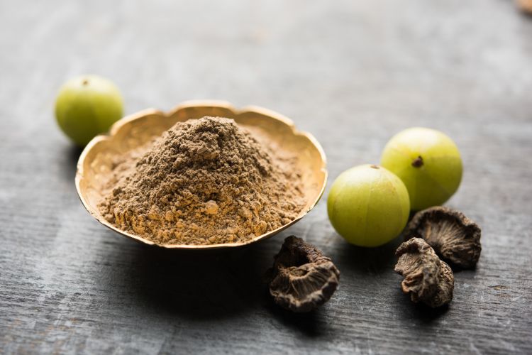 Amla: De Kleine Vrucht met Grote Gezondheidsvoordelen 3 Een gouden kom gevuld met bruin amlapoeder staat op een grijs houten oppervlak. Rondom de kom liggen drie verse groene amlas en drie gerimpelde gedroogde amlas. Het poeder is fijngemalen, wat contrasteert met de gladde en getextureerde oppervlakken van het verse en gedroogde fruit.
