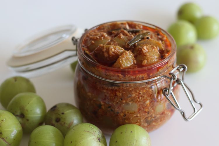 Amla: De Kleine Vrucht met Grote Gezondheidsvoordelen 4 Een glazen pot gevuld met Indiase kruisbessen augurk, met grove stukken en een rijke, gekruide mix. De pot heeft een scharnierend, luchtdicht deksel en is omgeven door hele, verse groene kruisbessen verspreid over een wit oppervlak. De augurk heeft een getextureerde en pittige uitstraling.