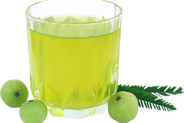 Amla: De Kleine Vrucht met Grote Gezondheidsvoordelen 5 Een glas gevuld met lichtgroen amla sap is omgeven door drie hele amla vruchten en twee groene bladeren. Het glas heeft een facet ontwerp en is geplaatst tegen een effen witte achtergrond. De levendige groene kleur contrasteert met het transparante glas en de textuur van de vruchten en bladeren.