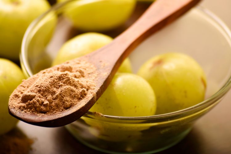 Amla: De Kleine Vrucht met Grote Gezondheidsvoordelen 6 Een glazen kom bevat verschillende hele, groene Indiase kruisbessen (amla). Een houten lepel rust op de kom, gevuld met bruinachtig amlapoeder. De achtergrond is zacht gefocust, waardoor de verse en gepoederde vormen van de vrucht worden benadrukt.