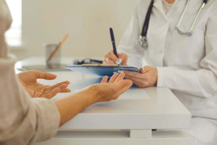 Hoofdpijn Na Yoga: Wat zijn de oorzaken en hoe kun je het voorkomen? 3 Een dokter in een witte jas zit aan een bureau en schrijft op een klembord. Aan de andere kant steekt een patiënt zijn handen uit in een vragend of verklarend gebaar. De scène suggereert een consult in een medische praktijk. Een beker met potloden is zichtbaar op het bureau.