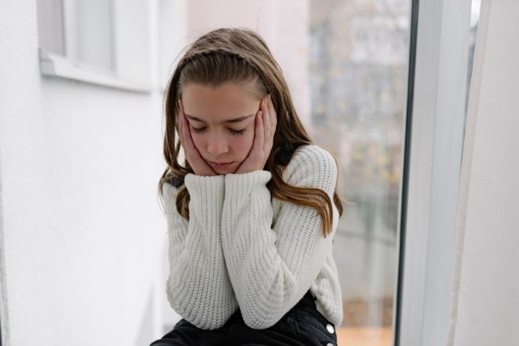Van Angst naar Succes: 7 Strategieën om je Kind te Helpen bij Optredens en Audities 1 Een jong meisje met lang bruin haar zit bij een raam, haar gezicht rustend in haar handen. Ze draagt een witte gebreide trui en lijkt peinzend of peinzend. De achtergrond is zacht wazig, waardoor een glimp van gebouwen buiten te zien is.