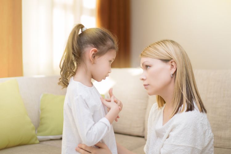 Van Angst naar Succes: 7 Strategieën om je Kind te Helpen bij Optredens en Audities 3 Een vrouw met blond haar knielt op de hoogte van een jong meisje met een paardenstaart. Ze zijn binnen, in een warm verlichte kamer met beige en groene kussens op een bank. De vrouw houdt de armen van het meisje zachtjes vast en beiden lijken verwikkeld in een serieus gesprek.