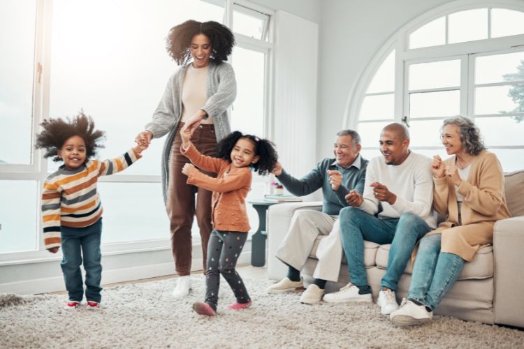 Van Angst naar Succes: 7 Strategieën om je Kind te Helpen bij Optredens en Audities 4 Een vrolijk familietafereel: twee volwassenen en drie kinderen dansen in een zonnige woonkamer met grote ramen. Een ouder echtpaar zit op een bank, lachend en klappend. De kinderen, een in strepen en de ander in oranje, zijn midden in de dans. De sfeer is warm en levendig.