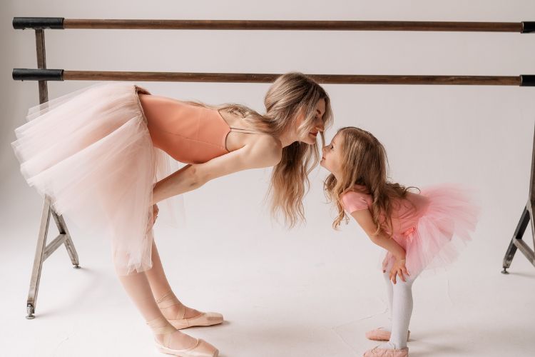 Van Angst naar Succes: 7 Strategieën om je Kind te Helpen bij Optredens en Audities 5 Een vrouw en een jong meisje, beiden in roze balletkleding, raken elkaars neuzen aan terwijl ze voorover buigen en naar elkaar lachen. Ze staan voor een balletbarre. De scène straalt warmte en vreugde uit, benadrukt door hun vergelijkbare outfits en sierlijke poses tegen een eenvoudige witte achtergrond.
