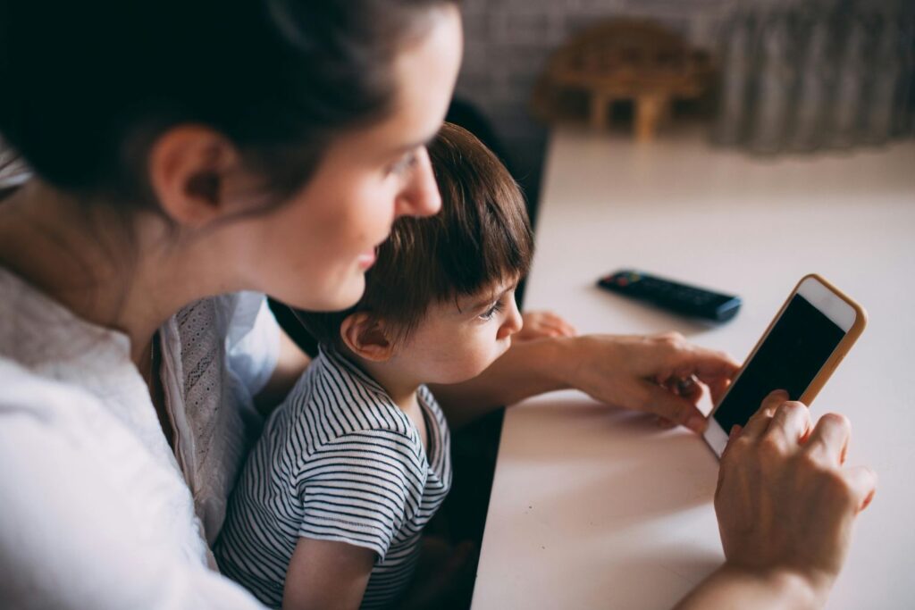 Een vrouw en een jong kind zitten aan tafel, beiden verdiept in een smartphone in de handen van de vrouw, de belichaming van de essentie van digitale opvoeding. Het kind, gekleed in een gestreept shirt, kijkt aandachtig toe. Een afstandsbediening ligt op tafel, terwijl een zacht wazige achtergrond hun op technologie gerichte betrokkenheid benadrukt.