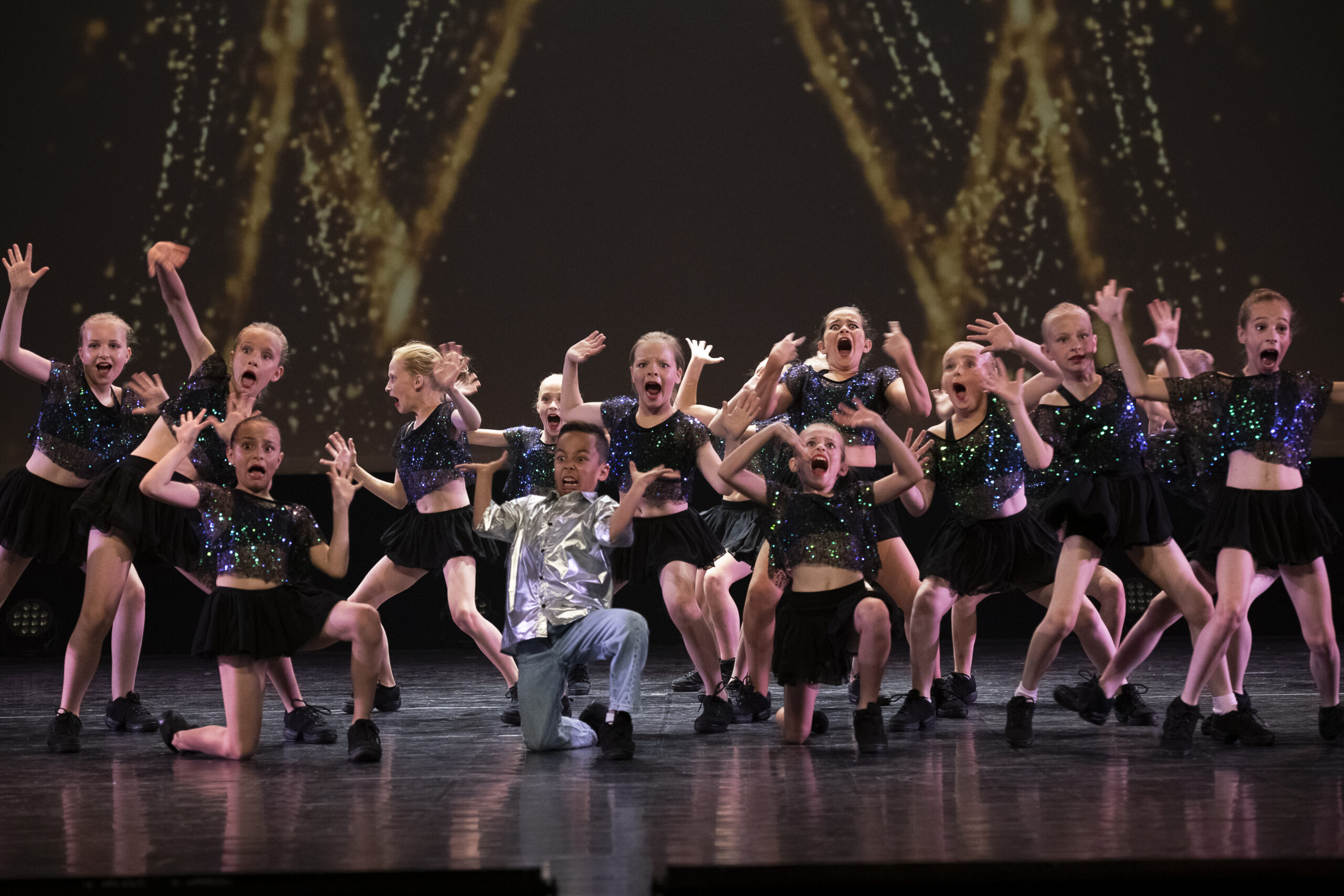 Jazz 1 Een groep jonge kinderen in glimmende, zwarte kostuums en één kind in een glimmend, zilveren shirt voeren een energieke dans uit op het podium. Ze trekken allemaal een expressief en verbaasd gezicht met hun handen omhoog, onder dramatisch gouden licht tegen een donkere achtergrond.