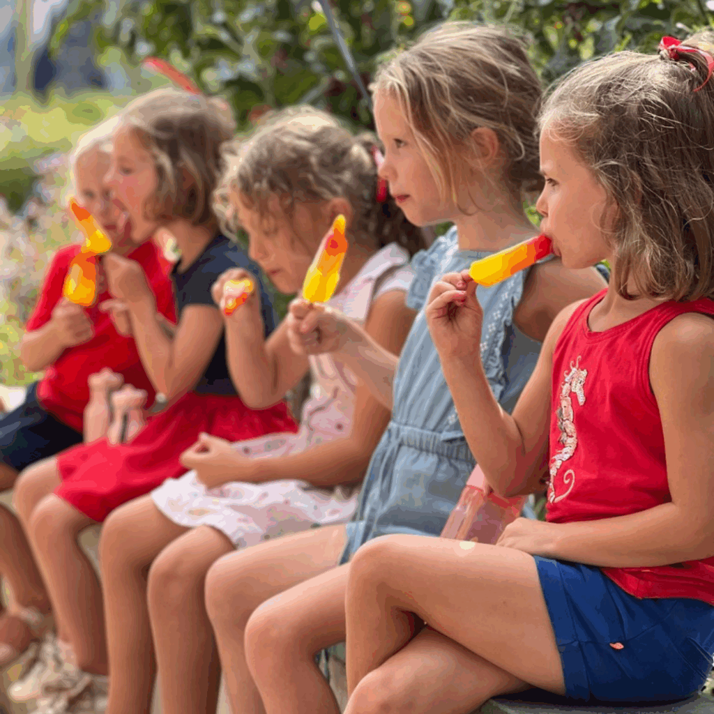 Vijf jonge meisjes zitten op een rij op een bankje buiten, elk genietend van een kleurrijk oranje-geel ijsje. Ze dragen zomerkleding in tinten rood, blauw en roze, met groen dat zacht vervaagd is op de achtergrond.