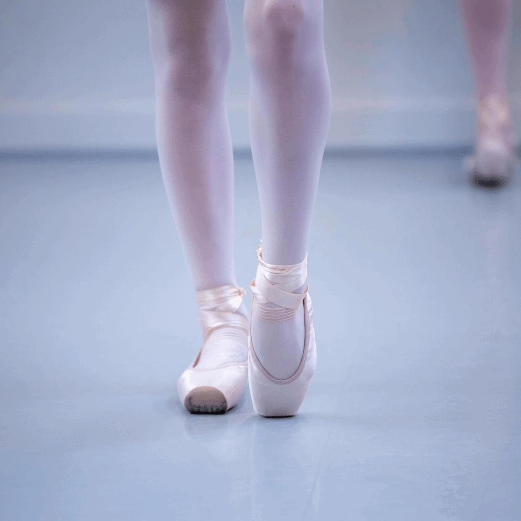 Een close-up van de benen van een balletdanseres toont haar staand op haar spitzen in roze balletschoenen en een witte panty op de lichtblauwe vloer van een dansstudio, met een andere danseres vaag op de achtergrond.