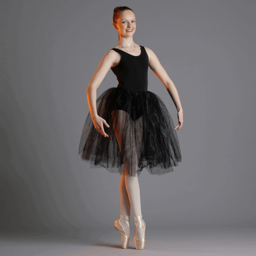 Een ballerina in een zwart turnpakje en een zwarte tule rok poseert op één voet en pointe, met haar armen gestrekt en omhoog, tegen een effen grijze achtergrond. Ze glimlacht en heeft haar haar netjes in een knotje.