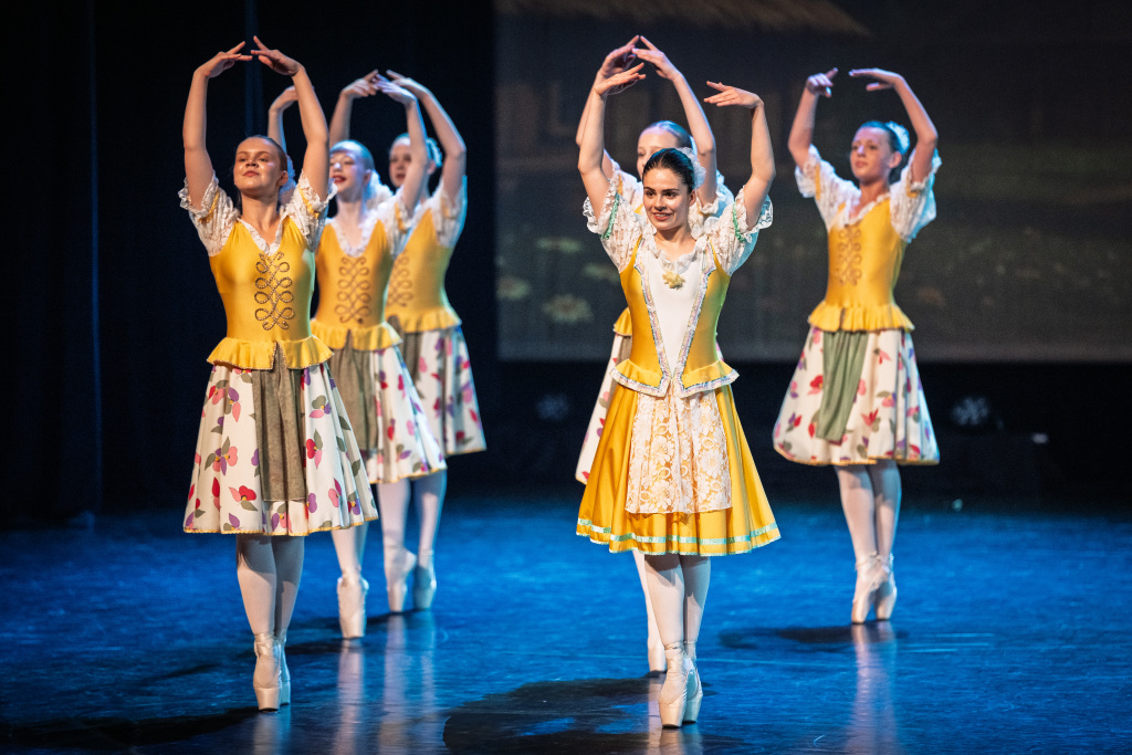 Vijf ballerina's in bijpassende gele en witte kostuums dansen en pointe op het podium, de armen sierlijk geheven, en voeren een klassiek repertoire uit onder zachte belichting.