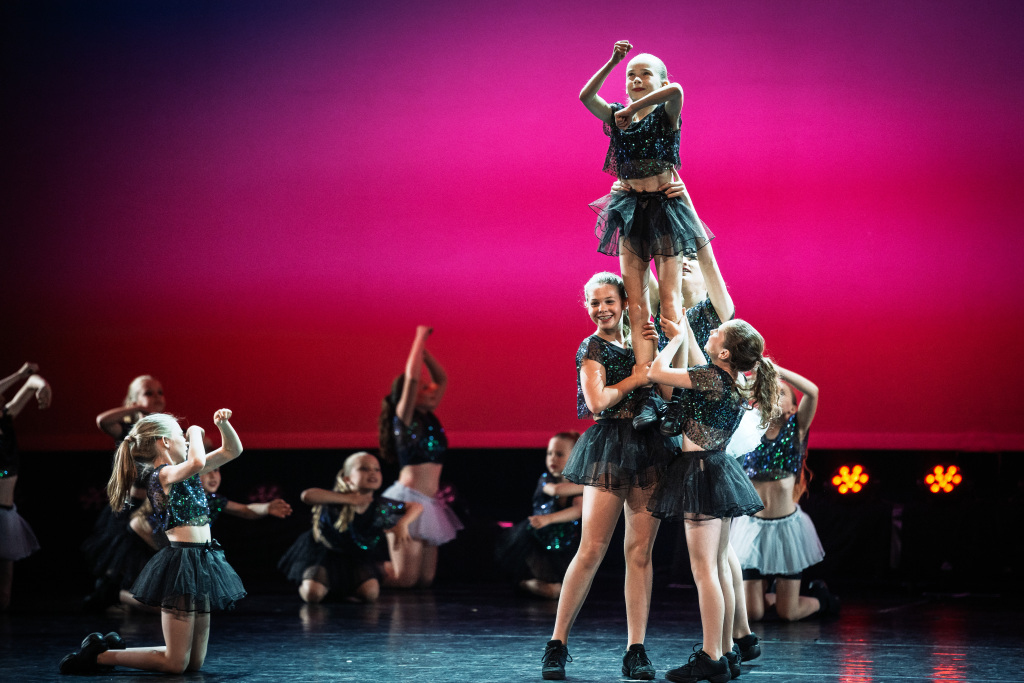 Een groep jonge dansers in zwarte glitterkostuums voert een funky dans uit en tilt een lachend meisje op het podium met een roze en paars verlichte achtergrond.
