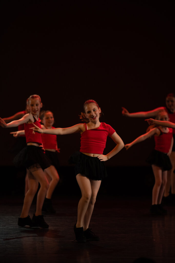 Een groep jonge meisjes voert een dansroutine uit op het podium. Het meisje vooraan lacht zelfverzekerd met één hand in haar zij. Alle dansers dragen bijpassende rode tops, zwarte rokken en zwarte schoenen, met donkere verlichting en een eenvoudige, donkere achtergrond.