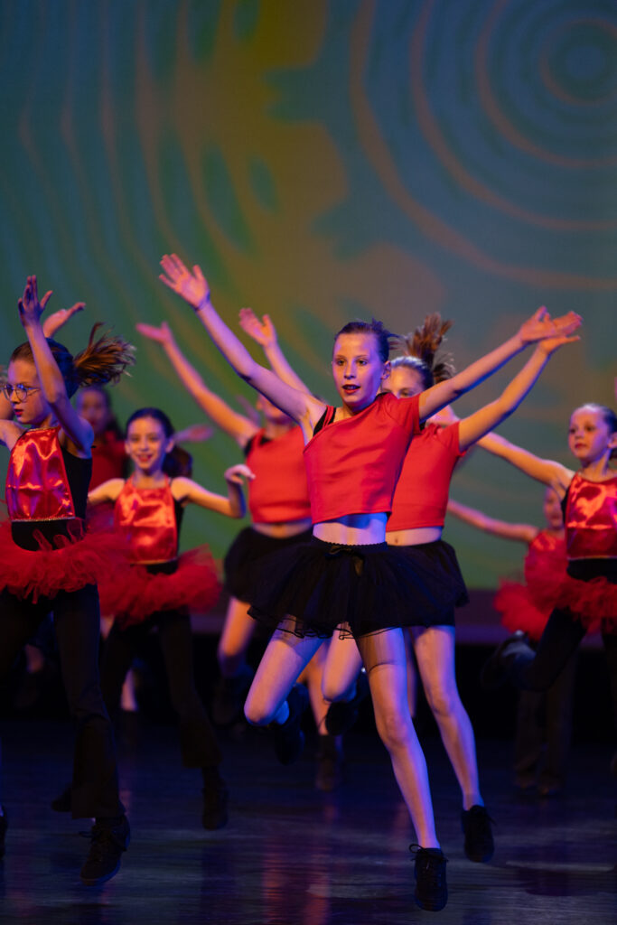 Een groep jonge meisjes in rode topjes en zwarte rokjes of shorts voert een energieke dans uit op het podium, met opgeheven armen. De achtergrond bestaat uit abstracte, wervelende gele en groene patronen en het podium is fel verlicht.