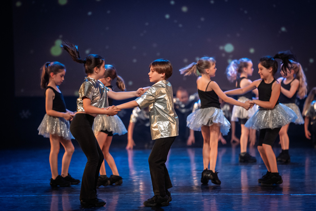 Kinderen in zilveren kostuums voeren paarsgewijs op het podium een levendige jazzdans uit, glimmende rokken vangen het licht tegen een blauwe achtergrond met sterren.