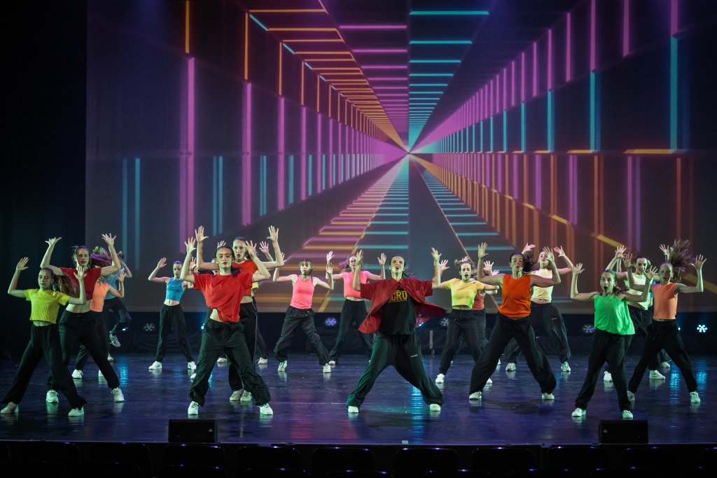 Hip Hop 1 Een grote groep dansers in kleurrijke shirts voert energieke Hip Hop uit op het podium, met een levendige geometrische tunnel geprojecteerd op de achtergrond.