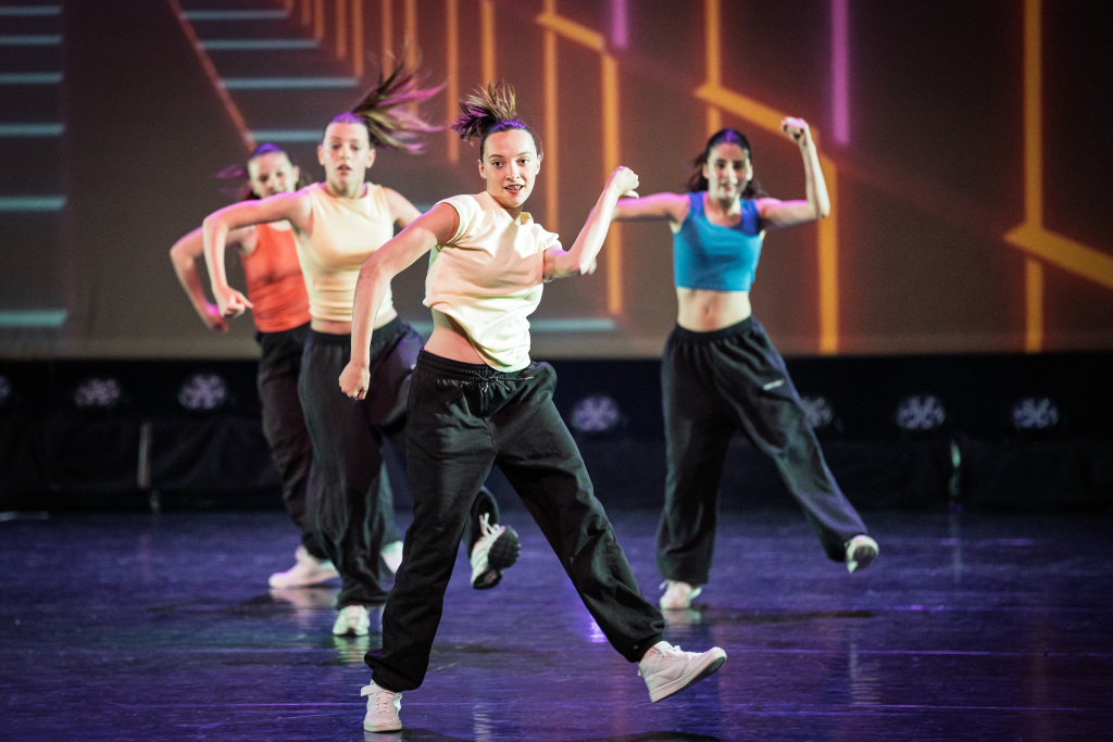 Hip Hop 7 Vier jonge vrouwen in sportieve outfits voeren een dynamische Hip Hop dansroutine uit op het podium, verlicht door levendige, kleurrijke lichten op de achtergrond.