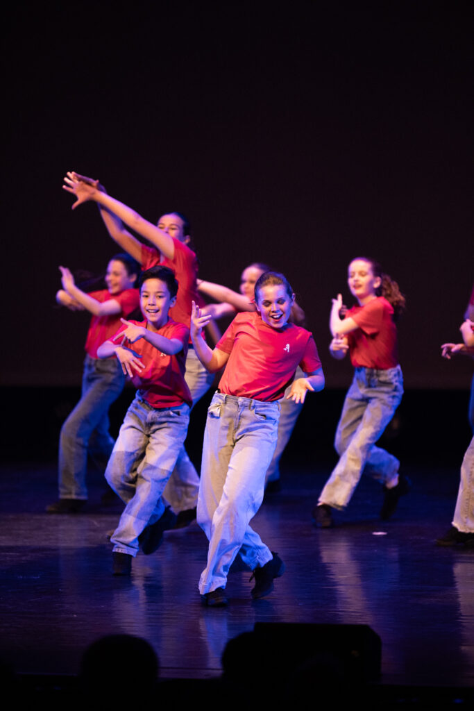 Een groep kinderen in rode shirts en blauwe spijkerbroeken voert een energieke dansroutine uit op het podium. Ze lachen en maken armgebaren, verlicht door de schijnwerpers op het podium en steken af tegen een donkere achtergrond.
