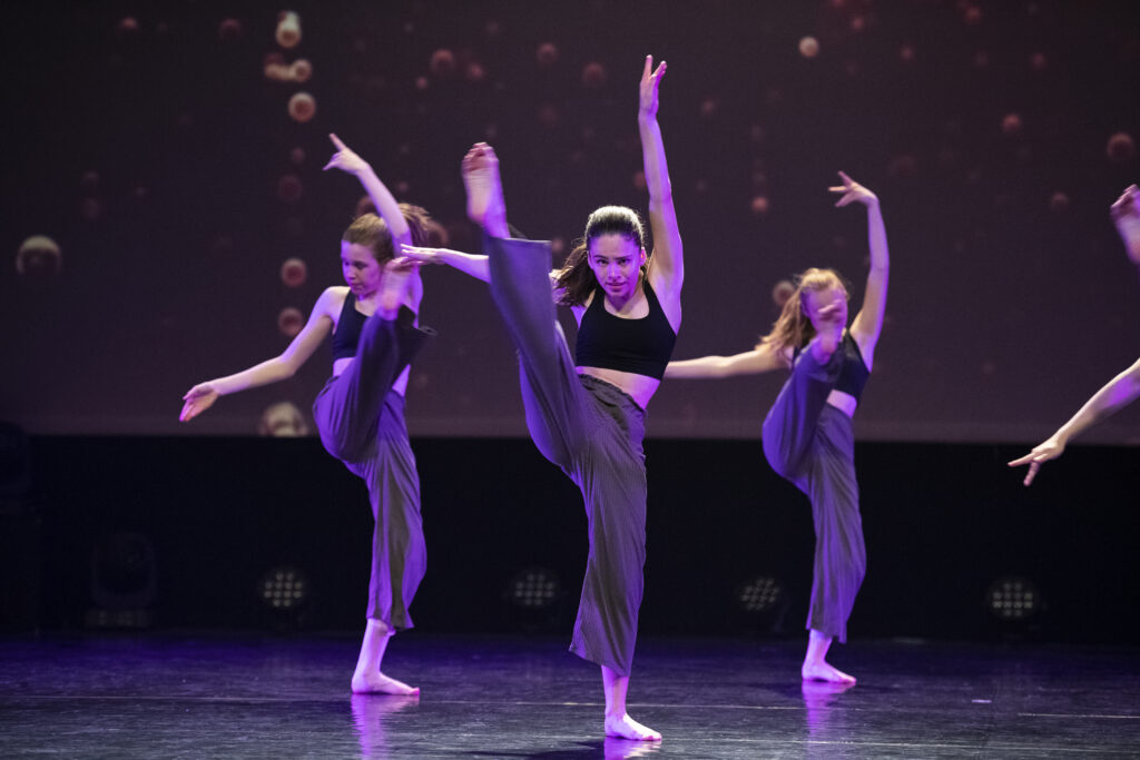 Hedendaagse dans 6 Drie danseressen treden op het podium op in donkere crop tops en losse broeken. De danseres in het midden balanceert op één been, haar andere been hoog gestrekt, haar armen omhoog. De achtergrond is donker met zacht licht. De sfeer is dynamisch en expressief.