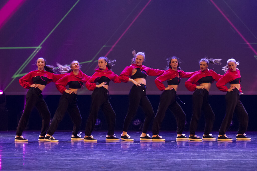 Acht dansers in bijpassende rode crop tops, zwarte high waist broeken en sneakers voeren een gesynchroniseerde hiphopdansroutine uit op het podium. Hun armen zijn gebogen, hun handen in hun zij, en hun haar zwiert, wat een dynamische beweging suggereert. Geometrische lichten schijnen achter hen.