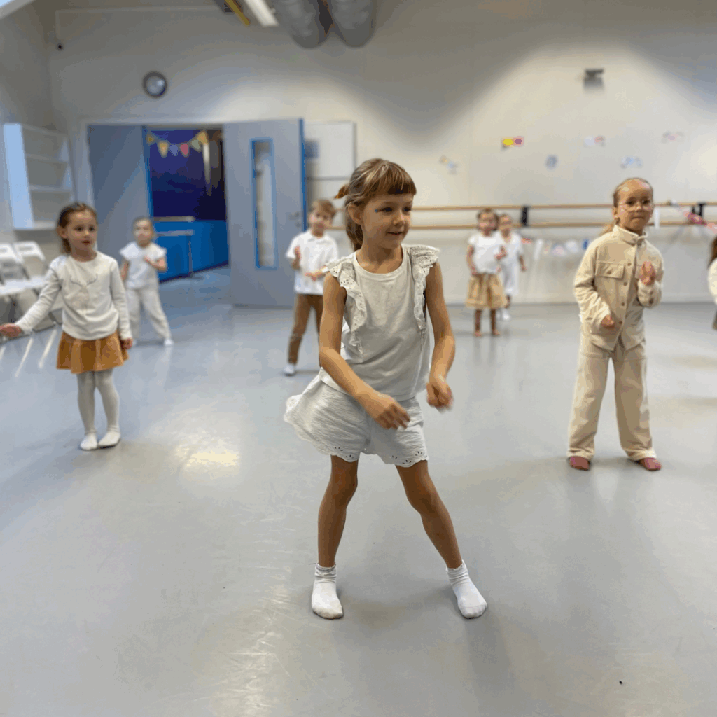 Een groep jonge kinderen, voornamelijk meisjes, staat verspreid in een lichte dansstudio. Het meisje vooraan draagt een witte jurk en sokken, midden in een dans. Andere kinderen in crèmekleurige of beige outfits staan achter haar. De kamer heeft een klok, slingers en balletstangen aan de muur.