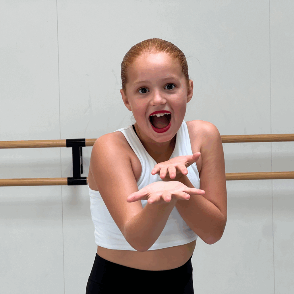 Een jong meisje met rood haar in een paardenstaart staat in een dansstudio, gekleed in een witte crop top en een zwarte legging. Ze heeft felrode lippenstift op, haar mond open van verbazing of opwinding, en haar handen voor zich uitgestrekt, met de handpalmen naar boven.