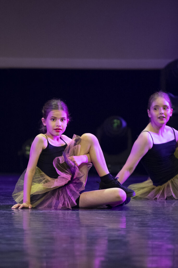 Twee jonge meisjes in bijpassende zwarte maillots en paarse tutu's zitten op een podiumvloer, midden in een dansvoorstelling. Het ene meisje zit met haar knie omhoog en haar hand op de vloer, terwijl het andere iets achter haar staat. De podiumverlichting zorgt voor een dramatisch effect.