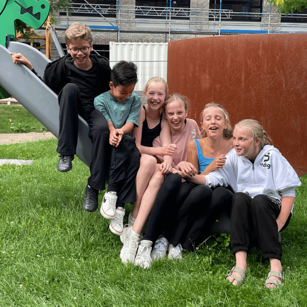 Zes lachende kinderen zitten dicht bij elkaar op een glijbaan in een park, lachend en elkaar vasthoudend. Ze dragen vrijetijdskleding en sneakers, omringd door groen gras. Op de achtergrond staan speeltoestellen en een bruine constructie.