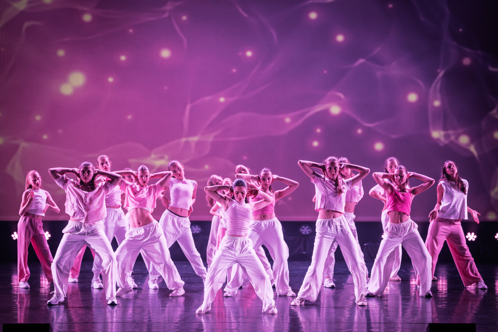 Girly Hip Hop 1 Een grote groep dansers in roze en witte outfits voert energieke Girly Hip Hop uit op het podium met een oplichtende paarse abstracte achtergrond en dramatische belichting.