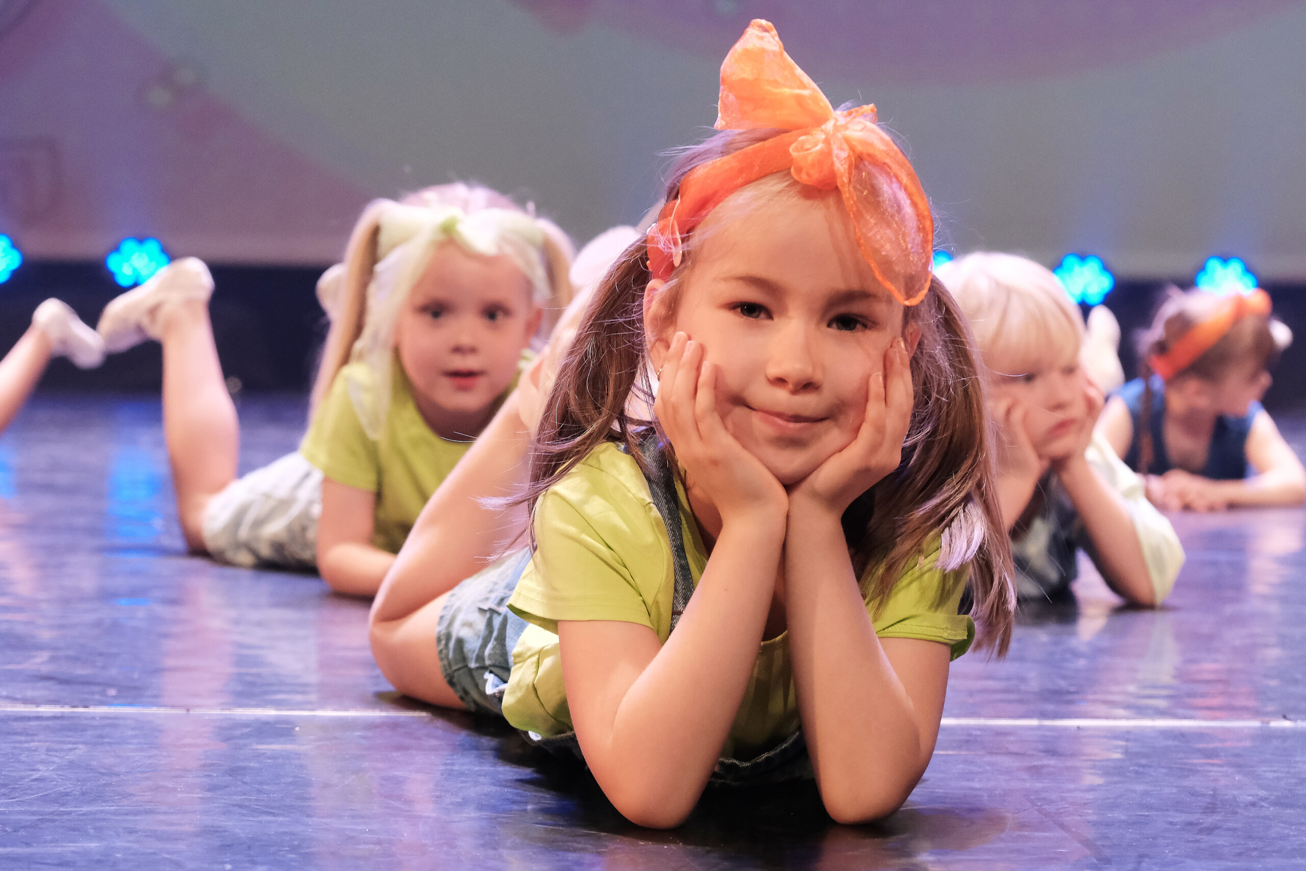 Kleuterdans 3 Jonge meisjes in felle outfits liggen glimlachend op het podium tijdens een vrolijk Kleuterdansoptreden, kleurrijke lichtjes schijnen op de achtergrond.