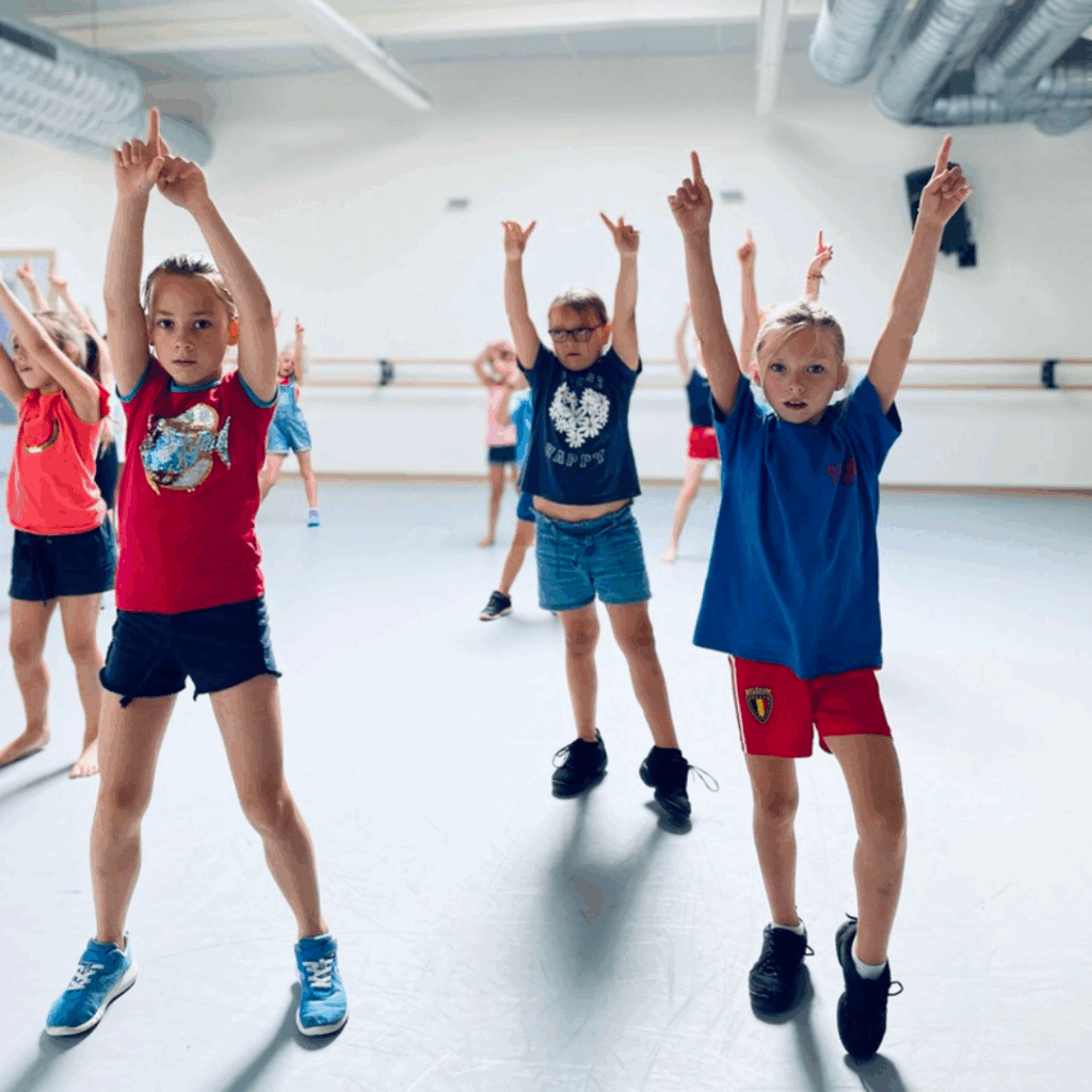 Een groep kinderen in casual sportkleding oefent een dansroutine in een lichte studio, waarbij ze één arm recht omhoog steken. Twee meisjes en een jongen op de eerste rij kijken geconcentreerd, met andere kinderen die erachter dansen. De ruimte heeft spiegels en balletstangen aan de muren.