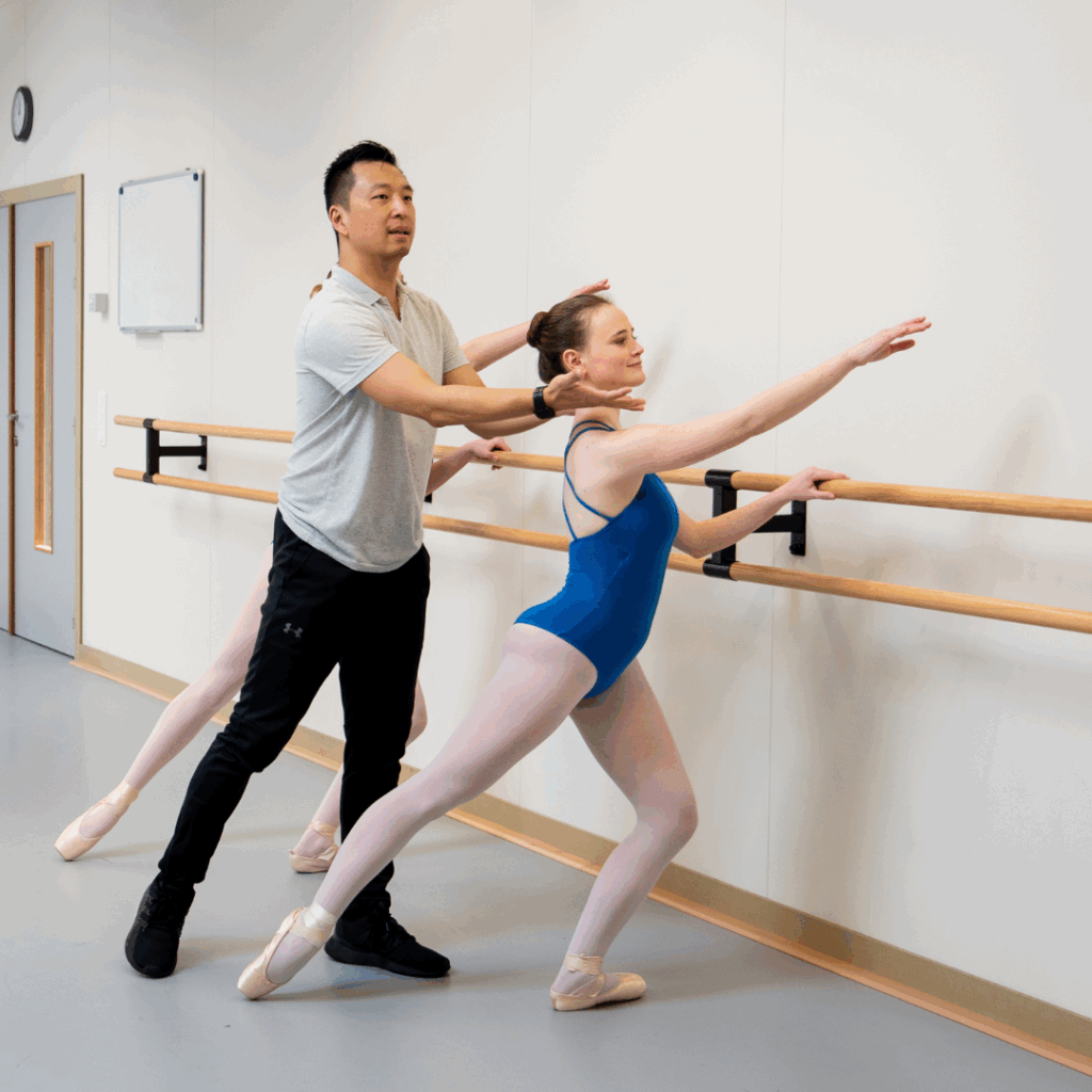 Private Coaching bij Ndigo 1 Een balletlerares in een grijs shirt begeleidt een danseres in een blauw balletpakje, roze maillot en spitzen terwijl ze oefent aan een balletbarre in een dansstudio. Private coaching sessie.