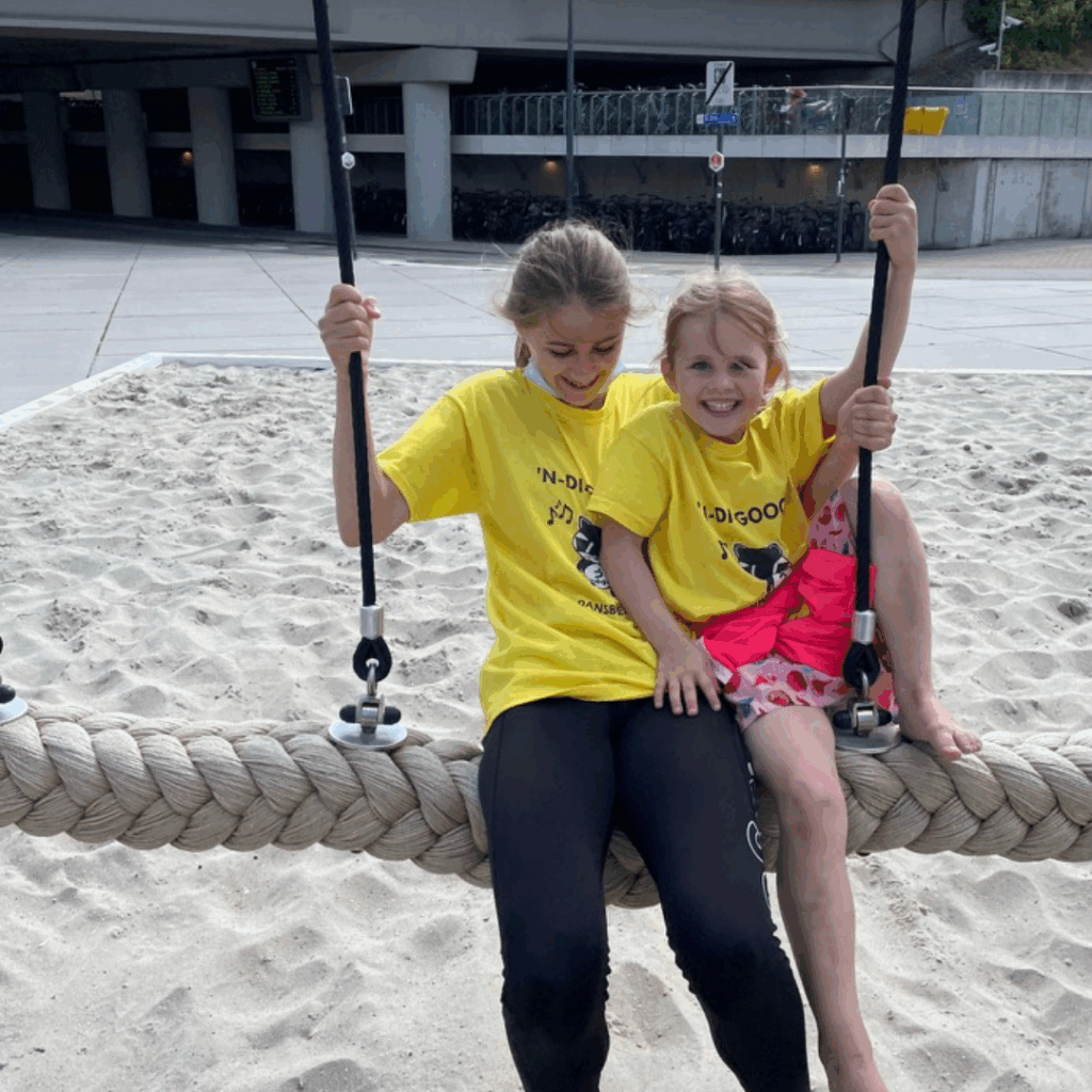 Twee lachende meisjes in gele T-shirts zitten samen op een grote touwschommel boven het zand. Het oudste meisje houdt de touwen vast, terwijl het jongste meisje op haar schoot zit met een arm om haar heen. Op de achtergrond zijn een viaduct en fietsen te zien.
