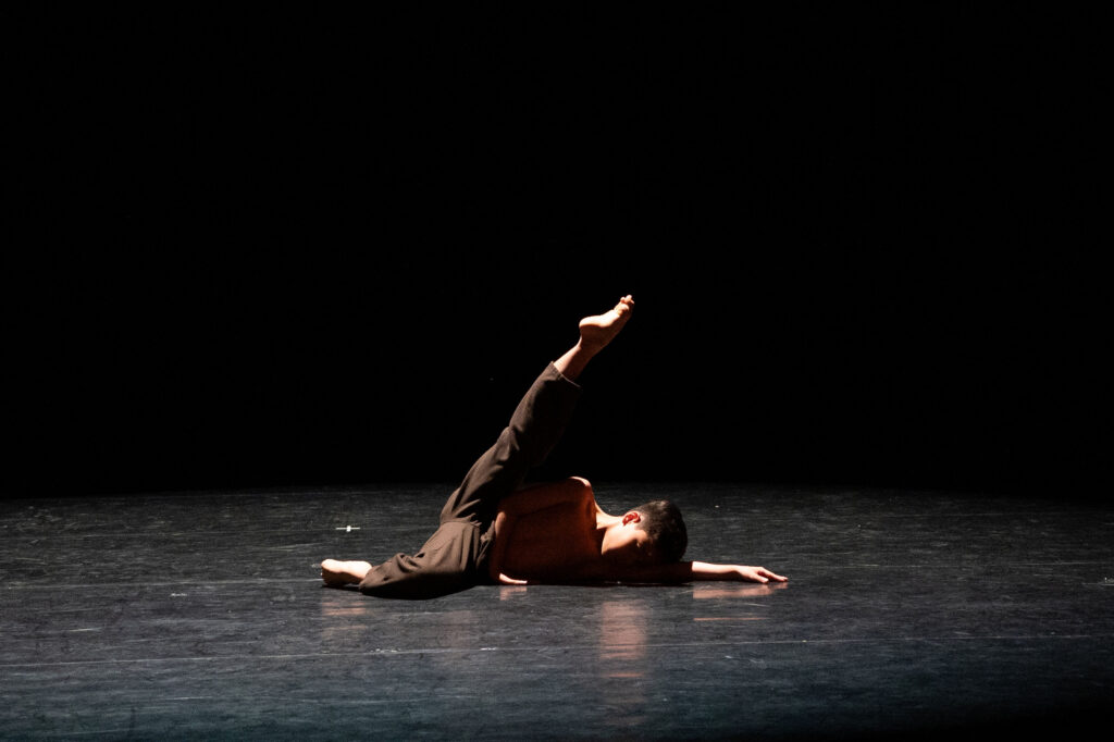 Een danseres ligt op een donker podium, één arm gestrekt en één been omhoog gestrekt. De danseres draagt een bruine broek en is shirtloos, beschenen door een spot tegen een zwarte achtergrond, wat een dramatische en minimalistische scène creëert.