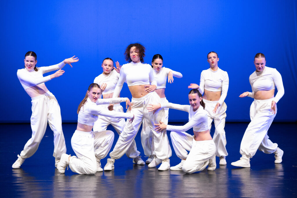 Girly Hip Hop 4 Acht dansers in witte outfits en sneakers treden op tegen een blauwe achtergrond. Ze poseren energiek met expressieve handgebaren en verschillende houdingen, en vormen een dynamische groepsopstelling, allemaal kijkend naar de camera.