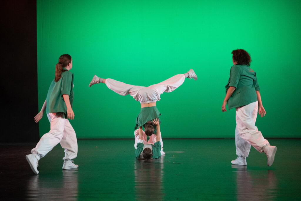 Drie dansers in groene shirts en witte broeken omringen een vierde danser die ondersteboven op zijn hoofd en handen balanceert, met zijn benen in een V-vorm. De achtergrond is effen felgroen en alle dansers dragen witte sneakers op een donker podium.