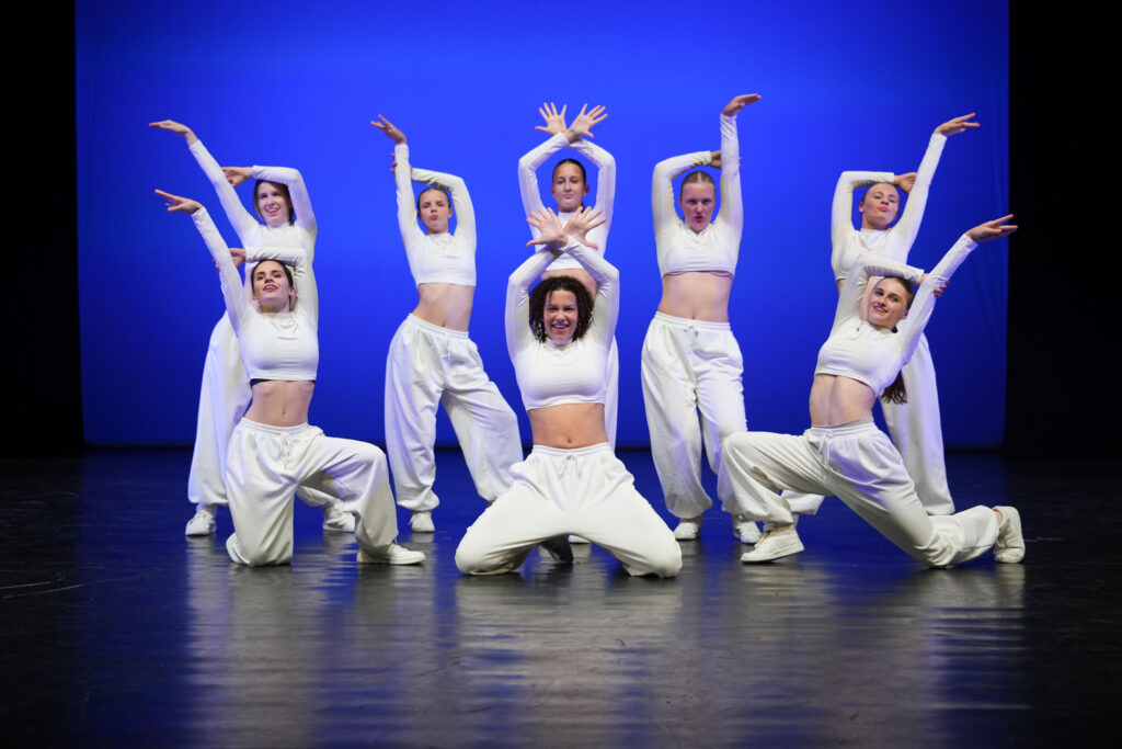 Girly Hip Hop 3 Acht dansers in witte crop tops, broeken en sneakers poseren in gecoördineerde, expressieve Hip Hop houdingen op een podium met een felblauwe achtergrond. Drie knielen vooraan, vijf staan erachter, allemaal met opgeheven of gebogen armen, wat een dynamische, symmetrische compositie creëert.
