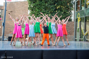 Een groep jonge dansers, waaronder een jazzdans groep, treedt op tijdens het optreden City Dance Fever. Gekleed in felle kleuren staan ze met opgeheven armen en gekruist, lachend op een buitenpodium tegen een stedelijke achtergrond van bomen en glazen gebouwen.