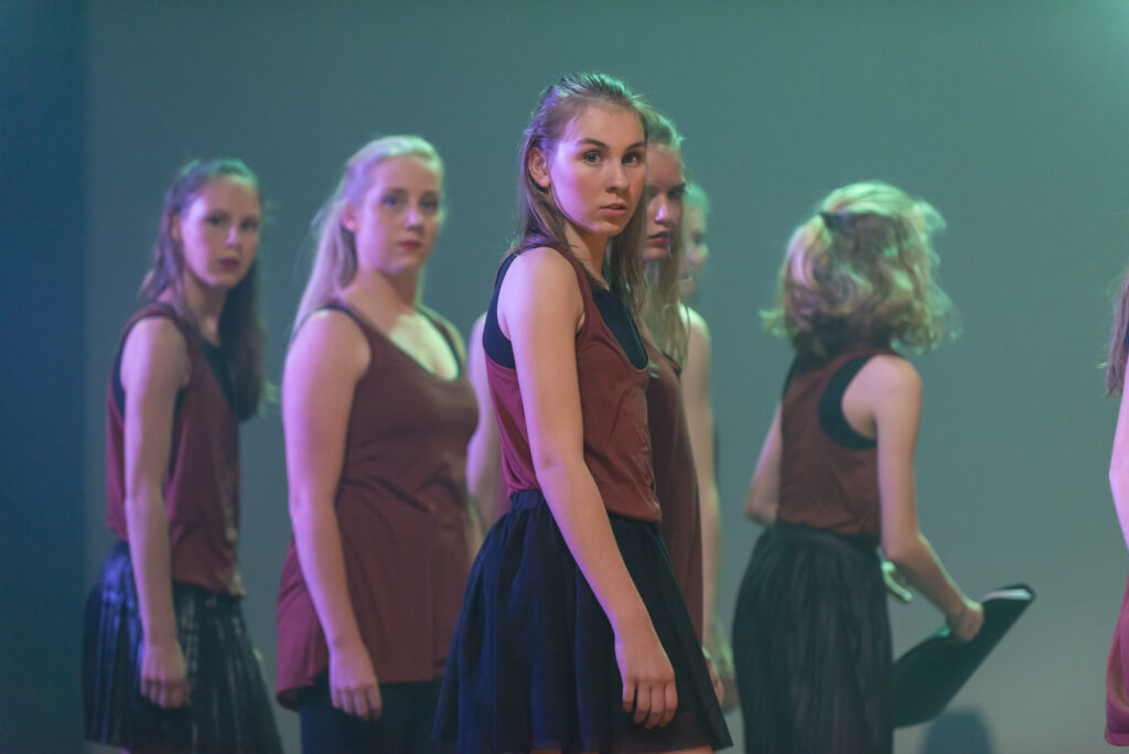 Hedendaagse dans 5 Een groep jonge vrouwen in bijpassende kastanjebruine topjes en zwarte rokken staat op het podium onder dramatische belichting. Ze lijken geconcentreerd en serieus, met één vrouw in het midden die recht in de camera kijkt, terwijl anderen in verschillende richtingen kijken.